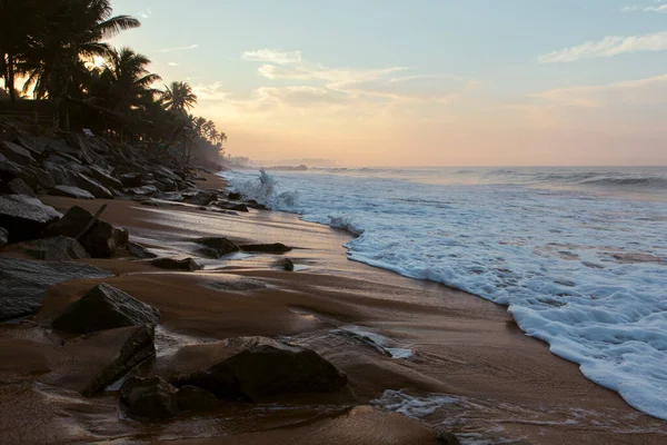 Pitiwella plajının (Sri Lanka) turuncu kum, yeşil palmiye ağaçları, kahverengi kayalar, büyük beyaz dalgalı Hint okyanusu ve ufukta güneş görünen güneş ile gün doğumunda çekilmiş güzel bir kartpostal manzarası..