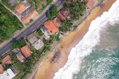 Deniz kıyısında bir yol, güzel villalar, yeşil ağaçlar, sarı kum, turkuaz mavi su, büyük dalgalar ve plaja düşen köpüklü hava aracı gündüz görüntüsü. Pitiwella, Sri Lanka