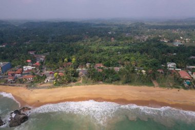 Deniz kıyısında bir yol, güzel villalar, yeşil ağaçlar, sarı kum, turkuaz mavi su, büyük dalgalar ve plaja düşen köpüklü hava aracı gündüz görüntüsü. Pitiwella, Sri Lanka