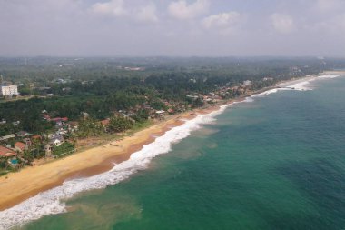 Deniz kıyısında bir yol, güzel villalar, yeşil ağaçlar, sarı kum, turkuaz mavi su, büyük dalgalar ve plaja düşen köpüklü hava aracı gündüz görüntüsü. Pitiwella, Sri Lanka