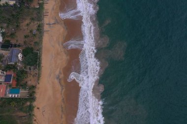 Deniz kıyısında bir yol, güzel villalar, yeşil ağaçlar, sarı kum, turkuaz mavi su, büyük dalgalar ve plaja düşen köpüklü hava aracı gündüz görüntüsü. Pitiwella, Sri Lanka