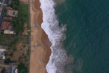 Deniz kıyısında bir yol, güzel villalar, yeşil ağaçlar, sarı kum, turkuaz mavi su, büyük dalgalar ve plaja düşen köpüklü hava aracı gündüz görüntüsü. Pitiwella, Sri Lanka