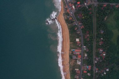Deniz kıyısında bir yol, güzel villalar, yeşil ağaçlar, sarı kum, turkuaz mavi su, büyük dalgalar ve plaja düşen köpüklü hava aracı gündüz görüntüsü. Pitiwella, Sri Lanka