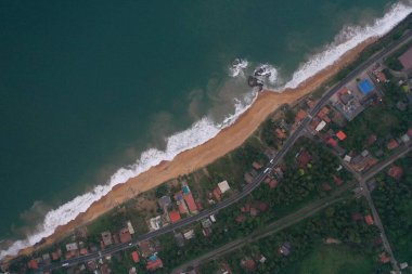 Deniz kıyısında bir yol, güzel villalar, yeşil ağaçlar, sarı kum, turkuaz mavi su, büyük dalgalar ve plaja düşen köpüklü hava aracı gündüz görüntüsü. Pitiwella, Sri Lanka