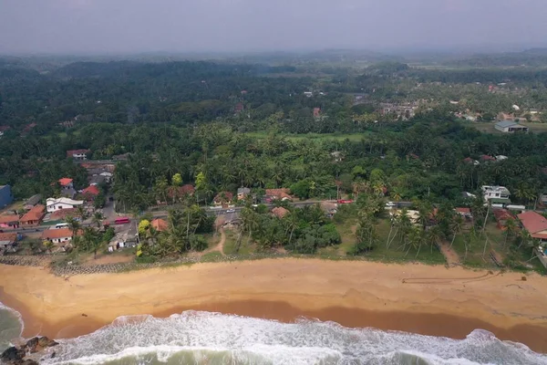 natural aerial drone bird view day shot of the sea shore with a road, beautiful villas, green trees, yellow sand, turquoise blue water, big waves and foam crashing on the beach. Pitiwella, Sri Lanka