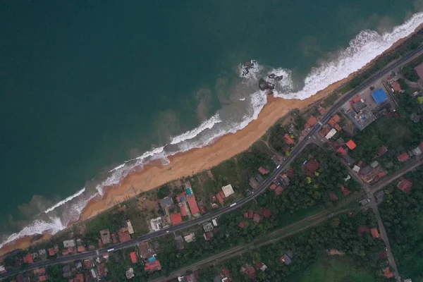 natural aerial drone bird view day shot of the sea shore with a road, beautiful villas, green trees, yellow sand, turquoise blue water, big waves and foam crashing on the beach. Pitiwella, Sri Lanka
