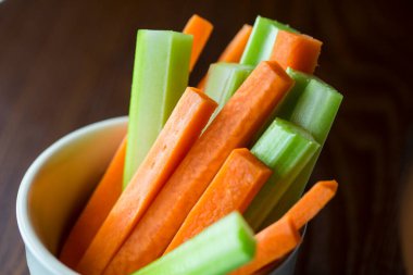 Çerçeveyi kapayın ve parti atıştırmalığının karesini çekin. Beyaz bir kase turuncu havuç ve sulu yeşil kereviz koyu kahverengi ahşap masa arkasında çubuklar.