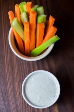Bir kase turuncu havuç ve yeşil sopa şeklinde koyu kahverengi ahşap bir masa üzerinde beyaz bir fincan mavi peynir sosu ile izole edilmiş üst görüntü görüntüsü.