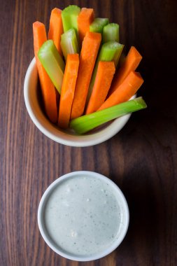 Bir kase turuncu havuç ve yeşil sopa şeklinde koyu kahverengi ahşap bir masa üzerinde beyaz bir fincan mavi peynir sosu ile izole edilmiş üst görüntü görüntüsü.