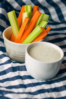 Partide atıştırmalık yiyeceklerin dikey yakın çekimi. Bir kase gevrek turuncu havuç ve sulu yeşil kereviz çubukları ile çizgili mavi ve beyaz havluya batırılmış beyaz peynir sosu.
