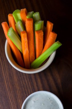 Bir kase turuncu havuç ve yeşil kereviz çubukları şeklinde koyu kahverengi ahşap bir masanın üzerinde beyaz bir fincan mavi peynirli sos ile izole edilmiş üst görüntü görüntüsü.