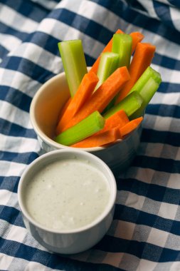 Partide atıştırmalık yiyeceklerin dikey yakın çekimi. Bir kase gevrek turuncu havuç ve sulu yeşil kereviz çubukları ile çizgili mavi ve beyaz havluya batırılmış beyaz peynir sosu.