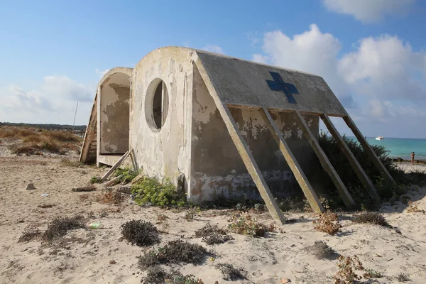 isolated abandoned concrete beach first aid station with a cross on the wall, standing in white sand with the ocean, blue sky,white clouds in the back. Shot on a summer day on Mallorca island, Spain