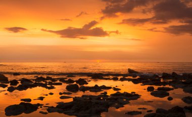 Dalgaların çekildiği kayalık bir sahilin zıt manzarası ve okyanus yüzeyinde güzel bir gökyüzü yansıması olan altın turuncu bir günbatımı. Koh Lanta, Tayland