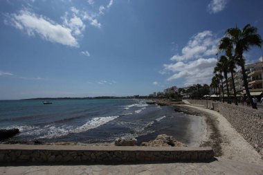 Yazın mavi deniz, beyaz dalgalar ve güzel bulutlu bir arka planda küçük bir tekne ile kıyı şeridinde kayaların ve uçurumların olduğu bir çekim. Cala Millor kasabası, Mallorca Adası, İspanya.