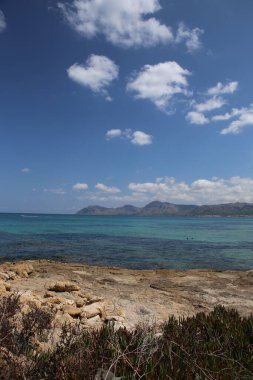 Panorama manzaralı ıssız kayalık plaj manzaralı mavi turkuaz deniz suyu ve beyaz bulutlu gökyüzü ve İspanya 'daki güzel ve renkli Mallorca adasında bir dağ arka planı
