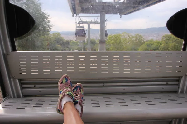 a close up shot of two crossed legs with pink camo sneakers and white socks on the feet stretched in the cabin of a funicular with trees and cable car cabins in the back. Barcelona, Spain