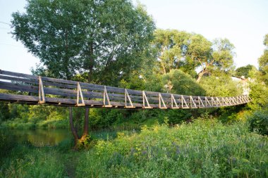 Güzel bir yaz günü, Nerl nehri üzerinde yüksek yeşil çimenler, canlı bitki örtüsü ve etrafındaki zengin yapraklı uzun ağaçlarla asılı eski bir ahşap köprü. Ivanovo, Vladimir Oblastı, Rusya