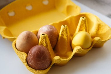 Sarı bir kutunun köşesinde tavuğa benzeyen dört farklı renkli ejderha veya dinozor doğal ürünlerle boyanmış Paskalya yumurtaları (kırmızı şarap, çay, havuç, zerdeçal).)