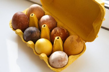 Sarı bir kutunun üst görüntüsünde sekiz farklı renkte ejderha ya da dinozor tavuk gibi görünüyor doğal ürünlerle boyanmış Paskalya yumurtaları (kırmızı şarap, çay, kahve, havuç, zerdeçal, soya sosu)