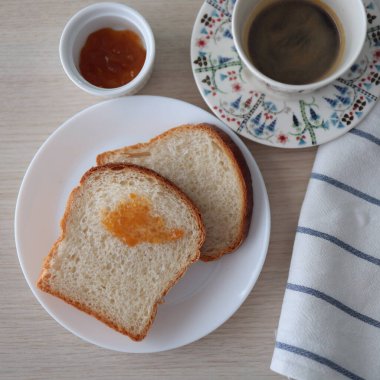 Beyaz tabakta kayısı reçelli sabah tostları ve bir fincan sade kahveyle servis ediliyor. Ahşap arkaplan ve üst görünüm.