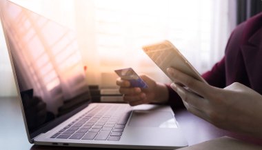 Kredi kartı tutuyor ve bilgisayarlı akıllı telefon kullanıyor. İnternetten alışveriş, e-ticaret, internet bankacılığı, harcama parası. Arka plan güneş ışığı kopyalama uzayı. Ev konseptinde online alışveriş.
