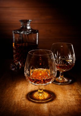 Kristal bir brendi bardağındaki spot ışığı. Arka planda bir sürahi, ahşap bir masanın üstünde çekilmiş..