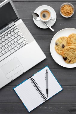 Çalışan bir kahve molasının portre görüntüsü. Laptop, kahve, kurabiye, şeker ve not defteriyle koyu ahşap bir arka planda.