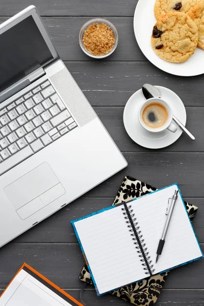 Çalışan bir kahve molasının portresi. Laptop, kahve, kurabiye, şeker ve koyu ahşap arka planda birden fazla not defteri.