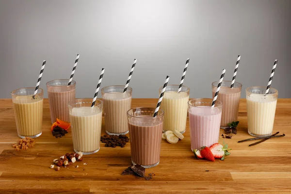 Glasses of various types of colourful milkshake with paper straws and ingredients on a wooden table top