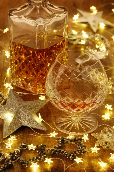 Crystal glass of brandy and a crystal brandy decanter with christmas lights and silver stars, on a wooden table