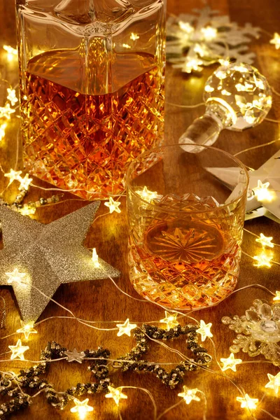 Crystal glass of whisky and a crystal whisky decanter with christmas lights and silver stars, on a wooden table