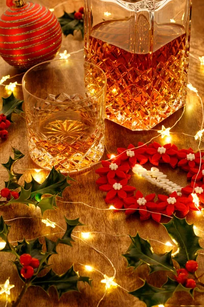 Crystal glass of whisky and a crystal whisky decanter with christmas lights and red christmas wreath decoration, on a wooden table