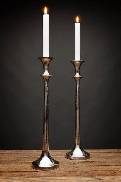 2 tall silver candle holders and glowing candles, shot on a wooden table, with a dark grey background