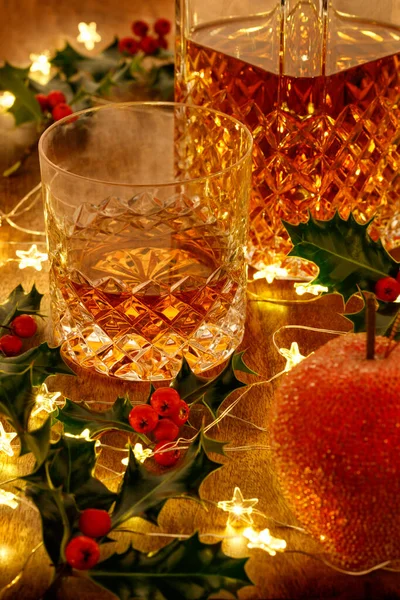 Kristal bardak viski ve noel ışıklandırmalı viski sürahisi ve ahşap bir masanın üzerinde kutsal böğürtlenler.