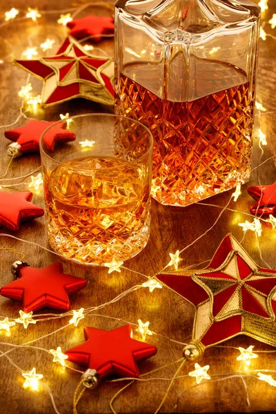 Kristal bardak viski ve noel ışıkları, takılar ve kırmızı yıldız süslemeleri olan bir viski sürahisi, ahşap bir masanın üzerinde.