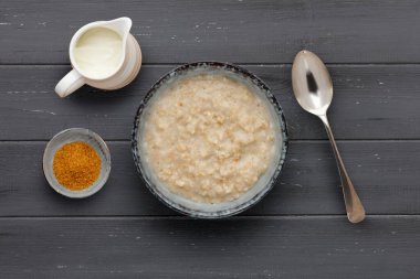 Besleyici bir kase yulaf lapası, küçük bir kase esmer şeker, bir sürahi süt ve bir kaşık, koyu ahşap arka planda.