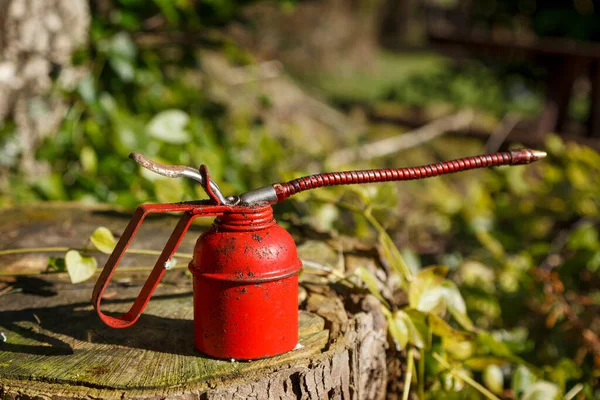 Old fashioned red oil can shot outside on a old tree stump, with green foliage in the background, on a sunny day.