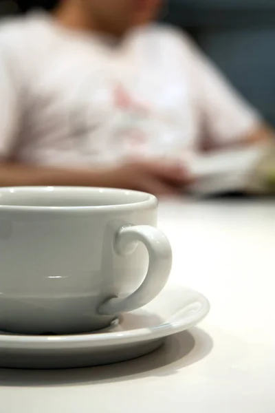 近照一张白色咖啡杯和茶托 放在白桌上 背后有一个注意力不集中的人在看书 — 图库照片