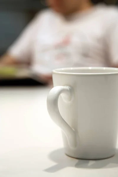 Close up shot of a white coffee mug, on a white table, with an out of focus person in the background, reading a book