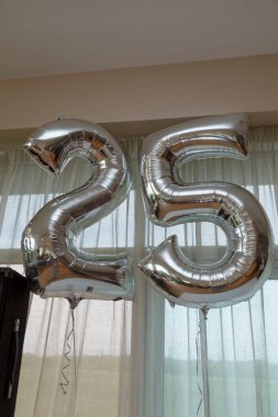 25 numaralı gümüş parti balonları, arka planda perdeler olan bir odada yüzüyor.
