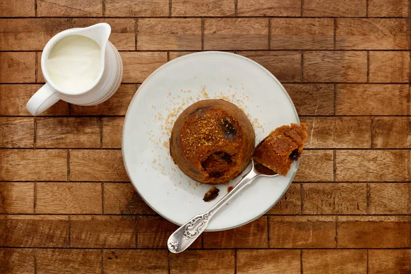 An individual spotted dick sponge cake, on a white plate with a spoon, and a small jug of cream, on a checked wooden surface with space for copy