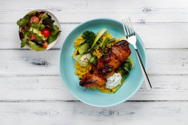 Lezzetli bir kase tandır tavuğu, sebzeler ve pilav, açık mavi bir kâse içinde, kırsal beyaz ahşap arka planda, yanında salata ile.