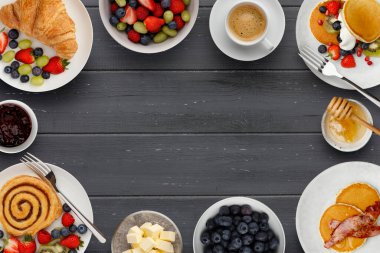 Lezzetli kahvaltı tabakları, krepler ve hamur işleri, bir kase meyve, reçel ve tereyağı ve bir fincan kahve, koyu ahşap arka planda fotokopi için yer var.