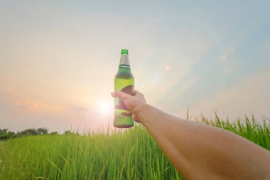 The young man holds a bottle of beer held in the evening sky on a beautiful Twilight light background above the green fields to congratulate the victory he has been drinking and celebrating beer alone. clipart