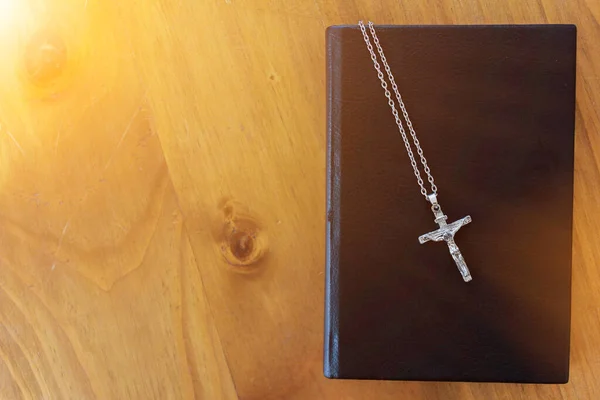A necklace with a pendant in the form of a holy cross was placed on the bible on a wooden table by the window in the morning. The cross and the Bible represent God that teaches love and forgiveness.
