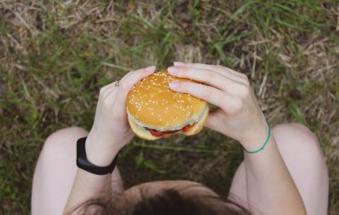 Kız çömelirken elinde bir hamburger tutuyor. Seçici odak, üst görünüm.