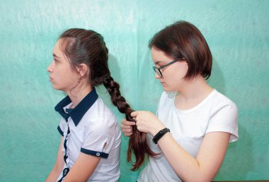 İki kız kardeş, büyük olan küçük kız kardeşinin saçını örüyor. Orta plan, turkuaz arkaplan.