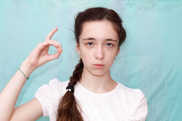 A girl with a long braid shows the letter D with her hand using the sign language of deaf-mutes. Medium plan, turquoise background.