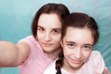 Gülümseyen iki güzel kız kardeş mavi arka planda akıllı telefonlarıyla selfie çekiyorlar. Kapat..
